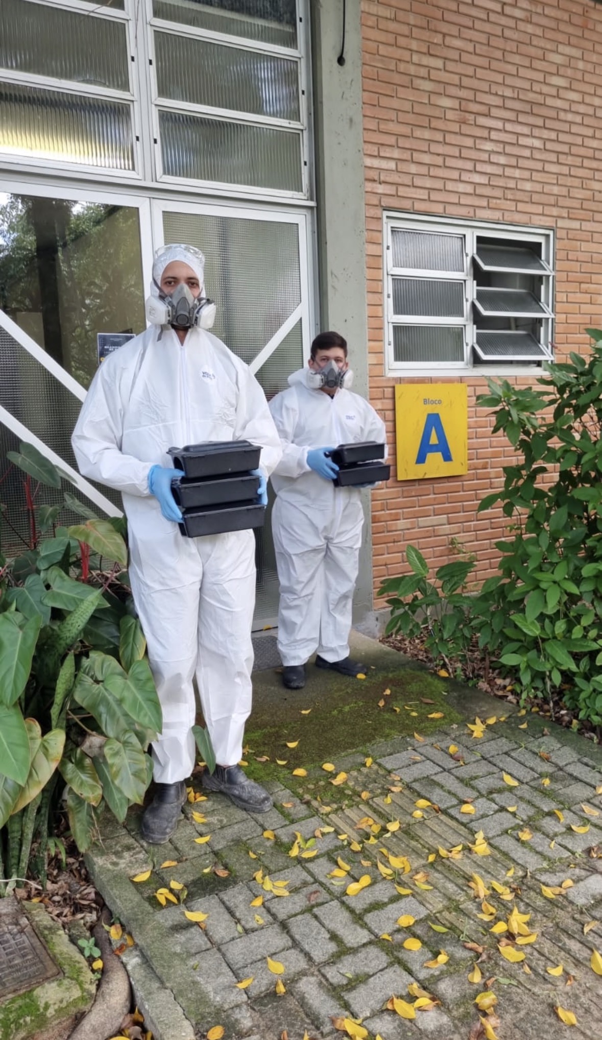 Dois técnicos NOVAECO uniformizados com macacão branco e EPIs carregando equipamentos de controle de cupins